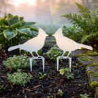 Sky white metal cardinal garden stakes placed in outdoor flower bed surrounded by greenery and stone garden edging