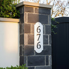 White vertical metal house number sign with cutout numbers mounted on a dark stone gate pillar