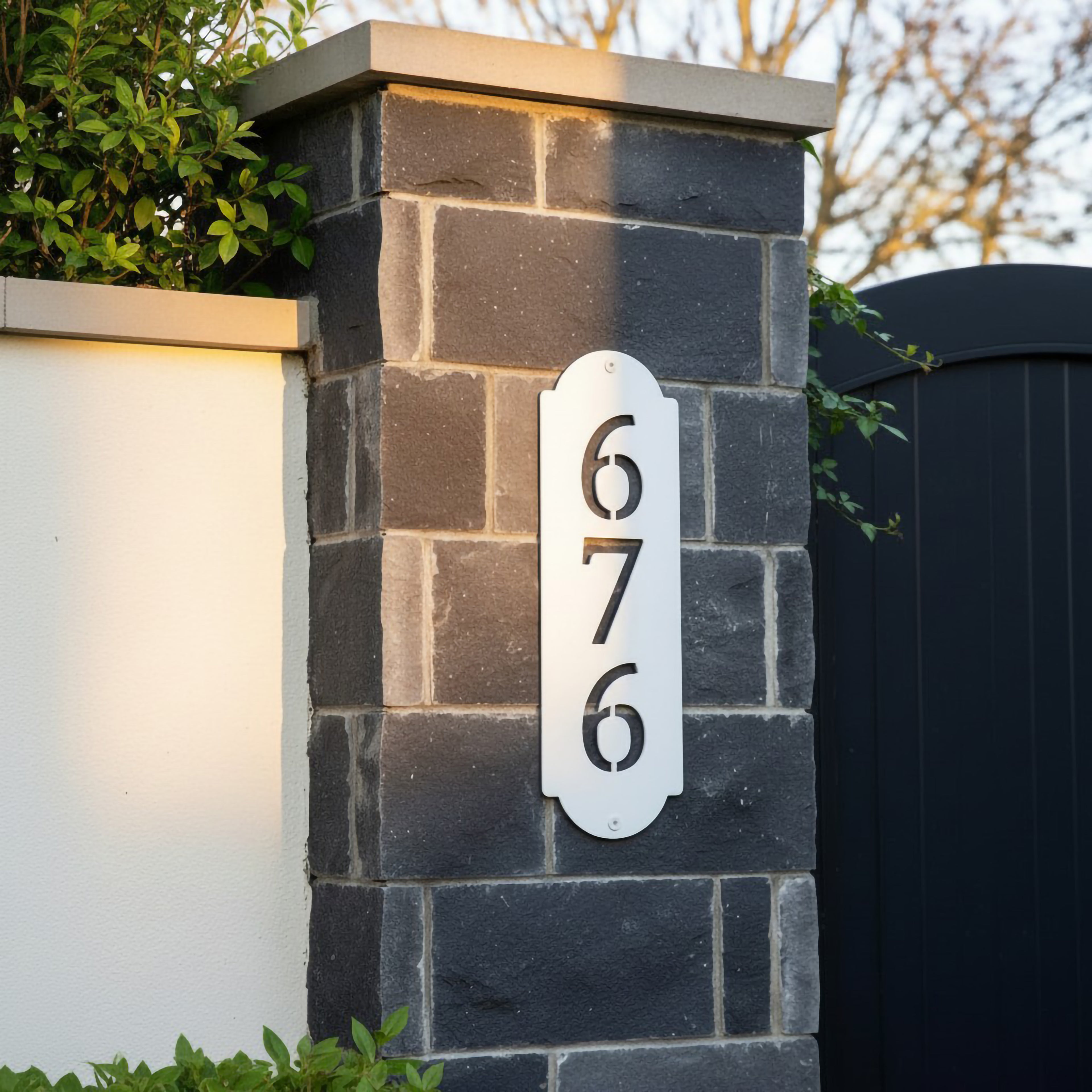 White vertical metal house number sign with cutout numbers mounted on a dark stone gate pillar