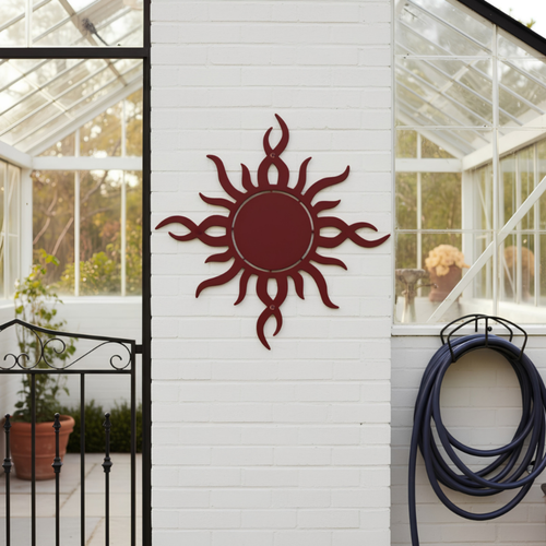 A deep red metal sunburst plaque with abstract wavy rays mounted centered on a white brick wall of a greenhouse, positioned between a black iron gate and a coiled blue garden hose.