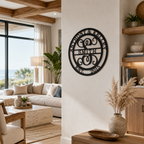 : A satin black circular metal monogram plaque reading "SMITH", "ANTHONY & KELLEY", and "EST. 2015" around an ornate script letter "S" is hung on a white pillar wall. The bright, open-concept coastal living room behind features a marble coffee table, beige couch, and tall glass windows overlooking the ocean.
