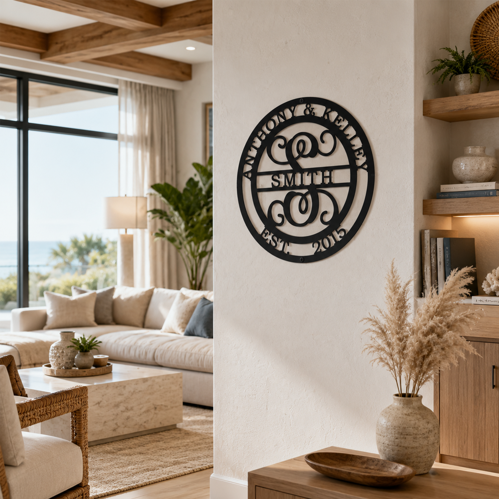 : A satin black circular metal monogram plaque reading "SMITH", "ANTHONY & KELLEY", and "EST. 2015" around an ornate script letter "S" is hung on a white pillar wall. The bright, open-concept coastal living room behind features a marble coffee table, beige couch, and tall glass windows overlooking the ocean.