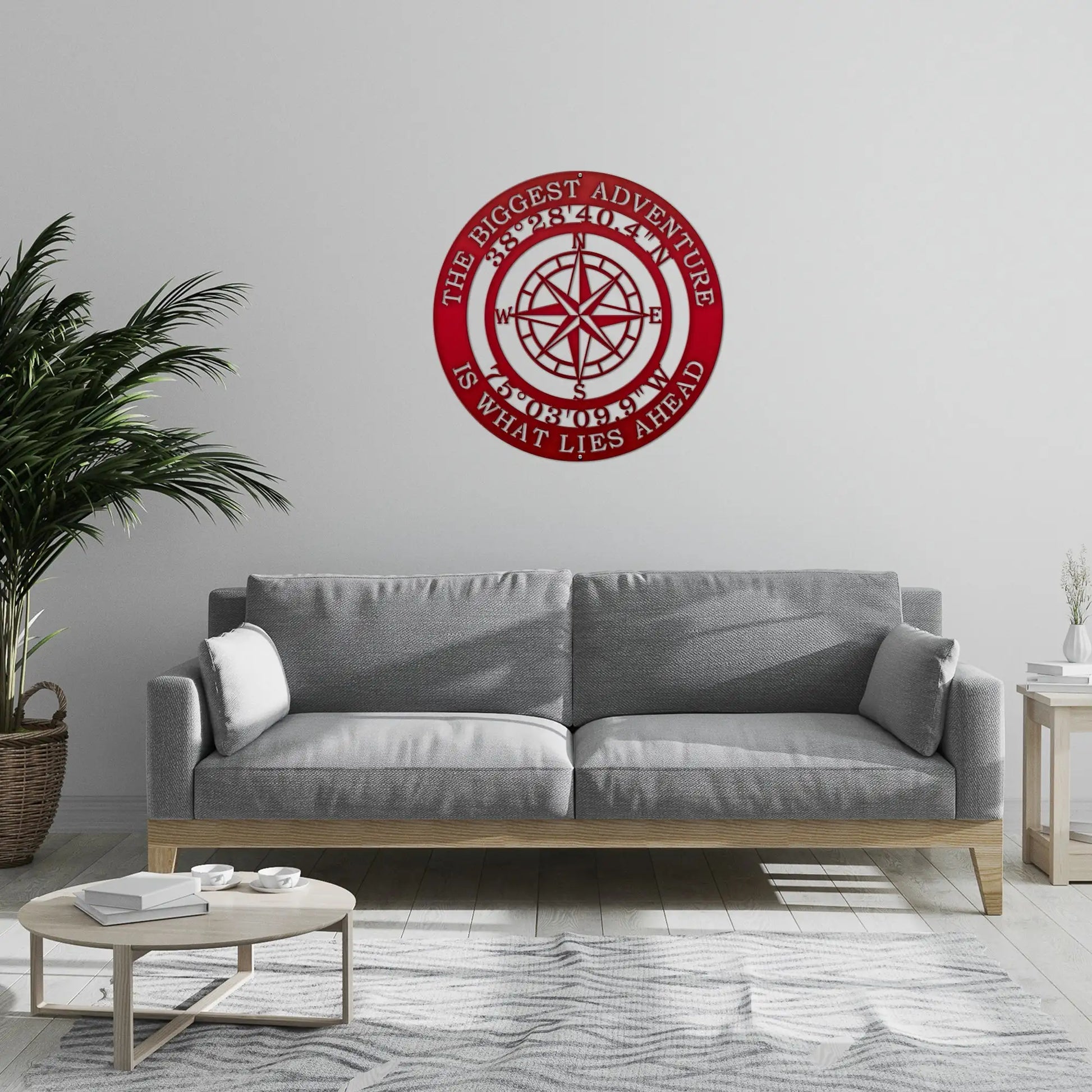 The Biggest Adventure Compass With Personalized GPS Coordinates, a metal sign featuring a compass design, is displayed leaning against a grey couch in a stylish living room setting.