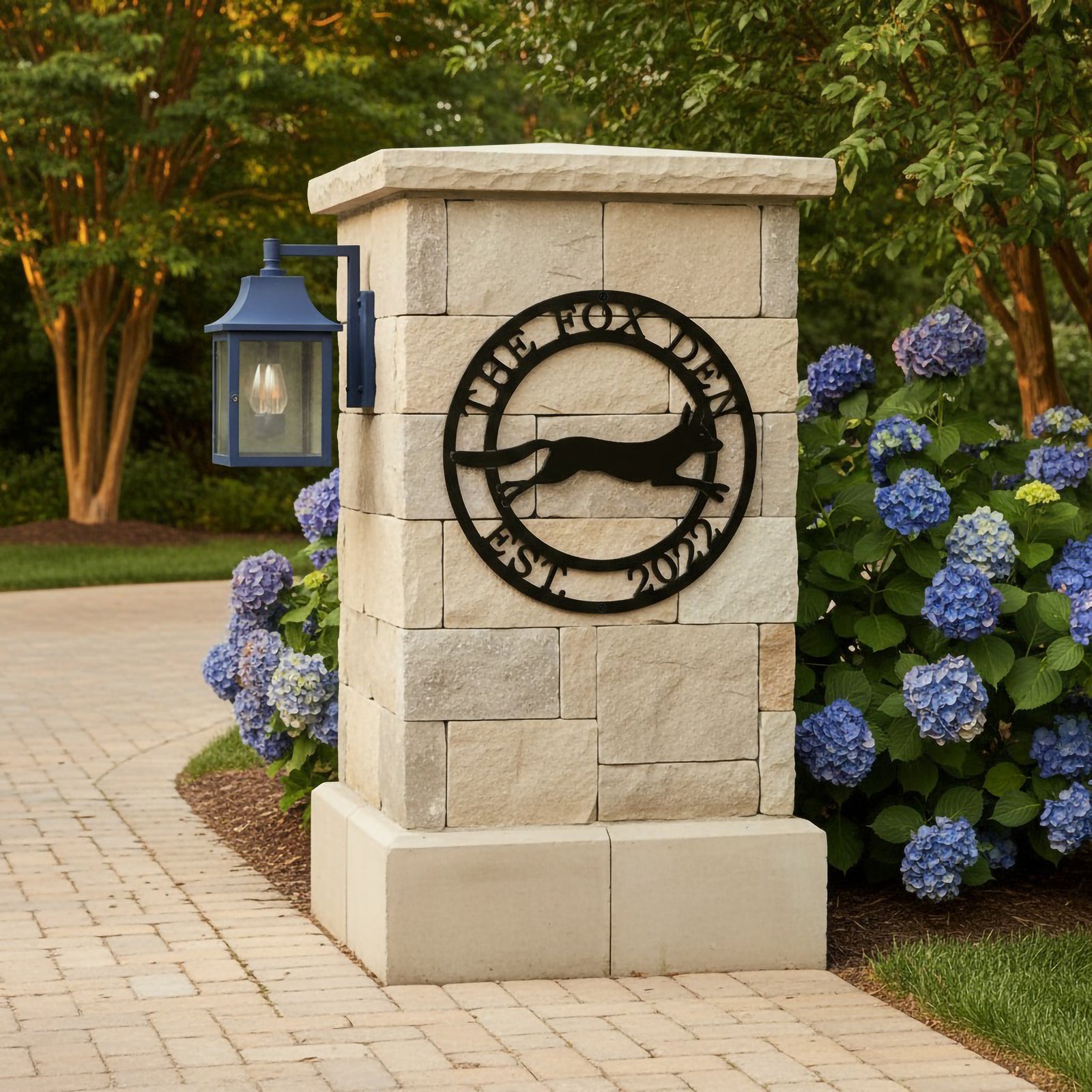 Black The Fox Den metal wall art with running fox design mounted on a stone pillar beside hydrangeas and outdoor pathway
