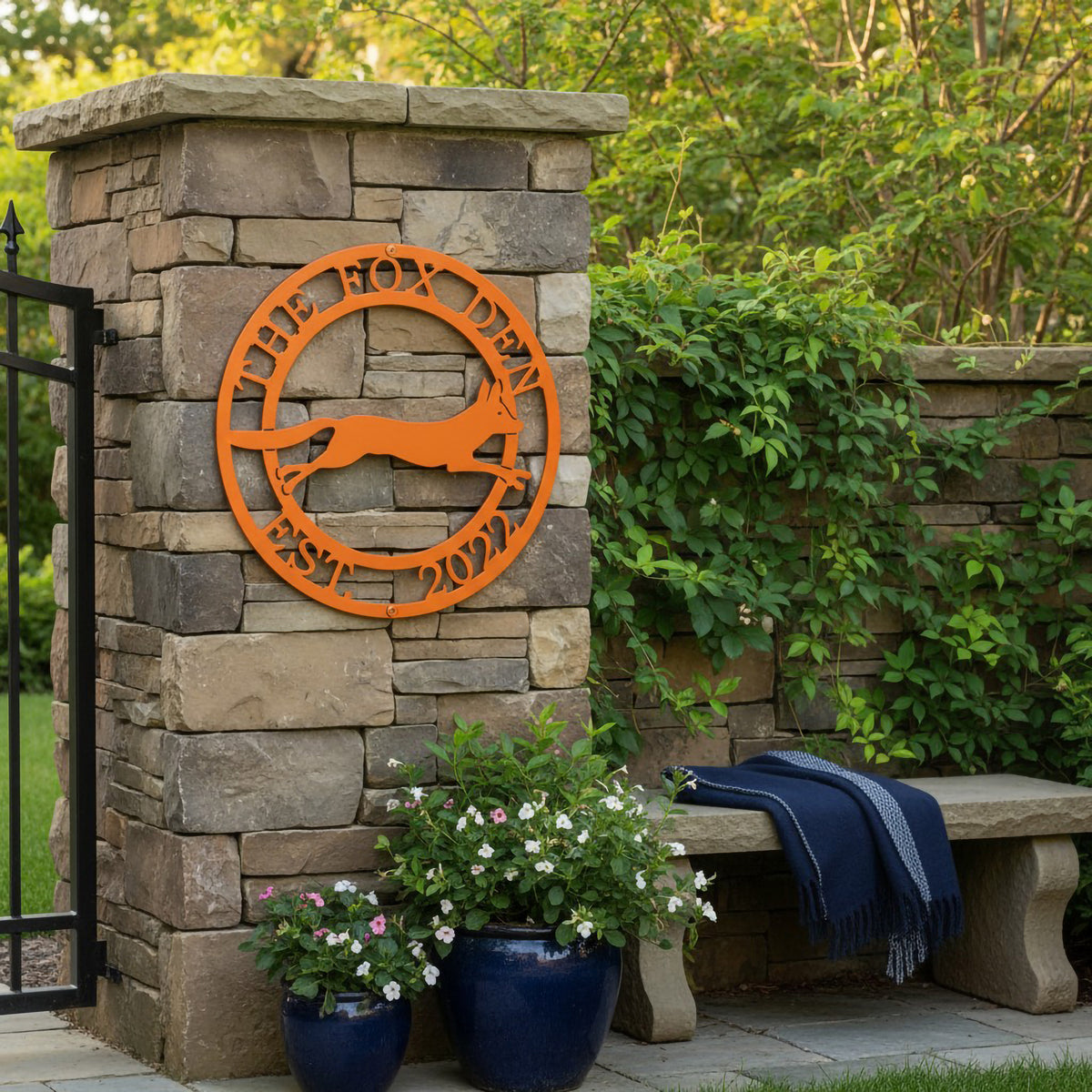 Orange The Fox Den metal wall art with fox silhouette displayed on a stone garden pillar with greenery and outdoor bench