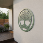 A circular metal sign with a muted sage green finish displaying an intricate Tree of Life with swirling branches. The text "THE GRANTHAMS" arches across the top border, and "EST. 1992" anchors the bottom. The plaque is mounted on a plain white exterior wall next to an outdoor porch with potted lavender plants.