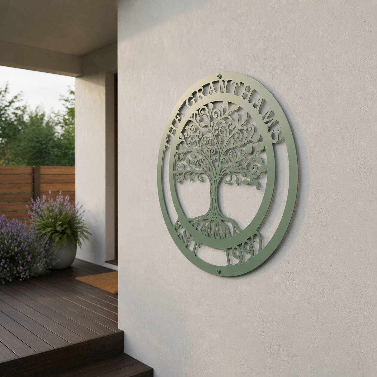 A circular metal sign with a muted sage green finish displaying an intricate Tree of Life with swirling branches. The text "THE GRANTHAMS" arches across the top border, and "EST. 1992" anchors the bottom. The plaque is mounted on a plain white exterior wall next to an outdoor porch with potted lavender plants.