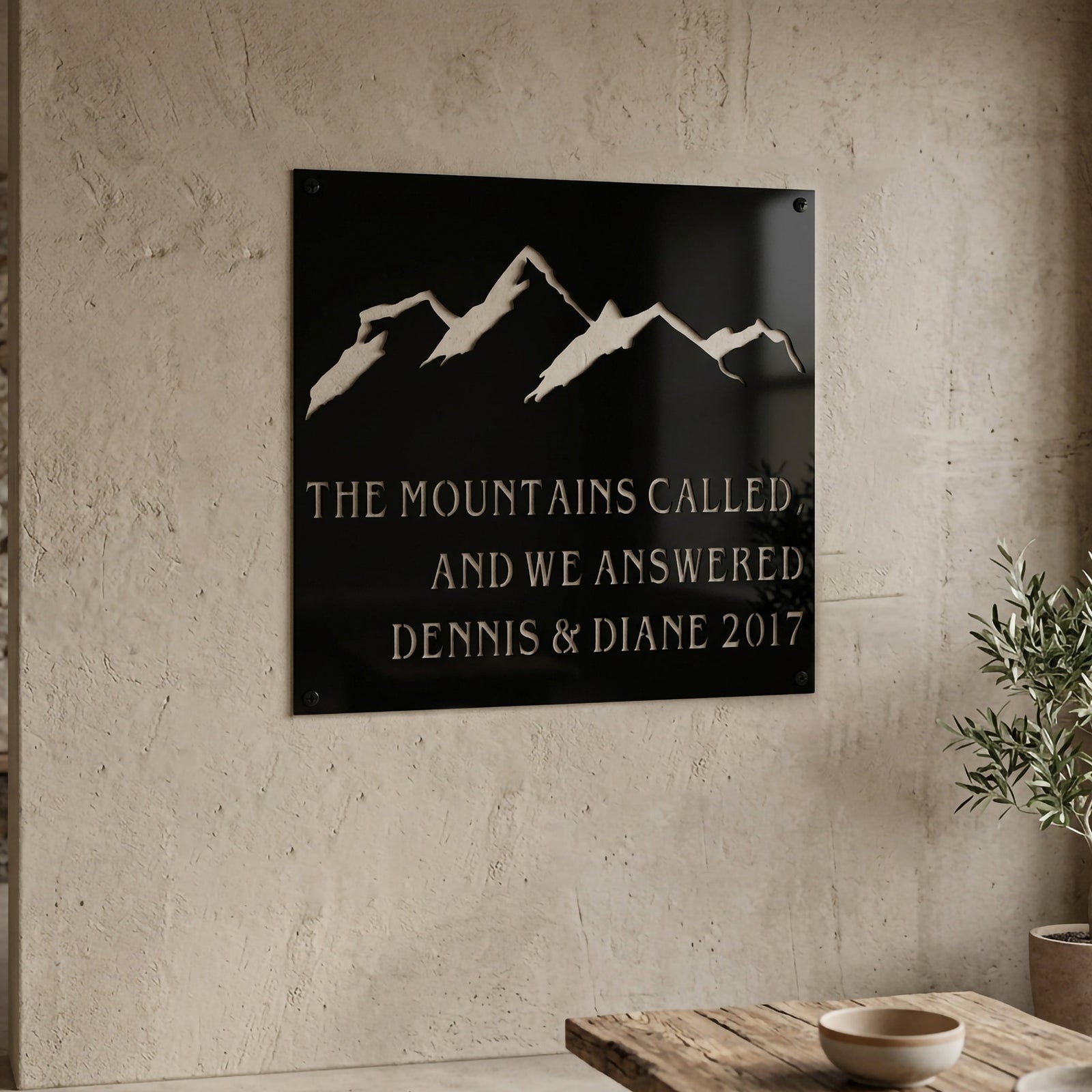 Emerald green mountain metal sign with personalized family names and established year displayed on an outdoor garden wall surrounded by plants and flowers.