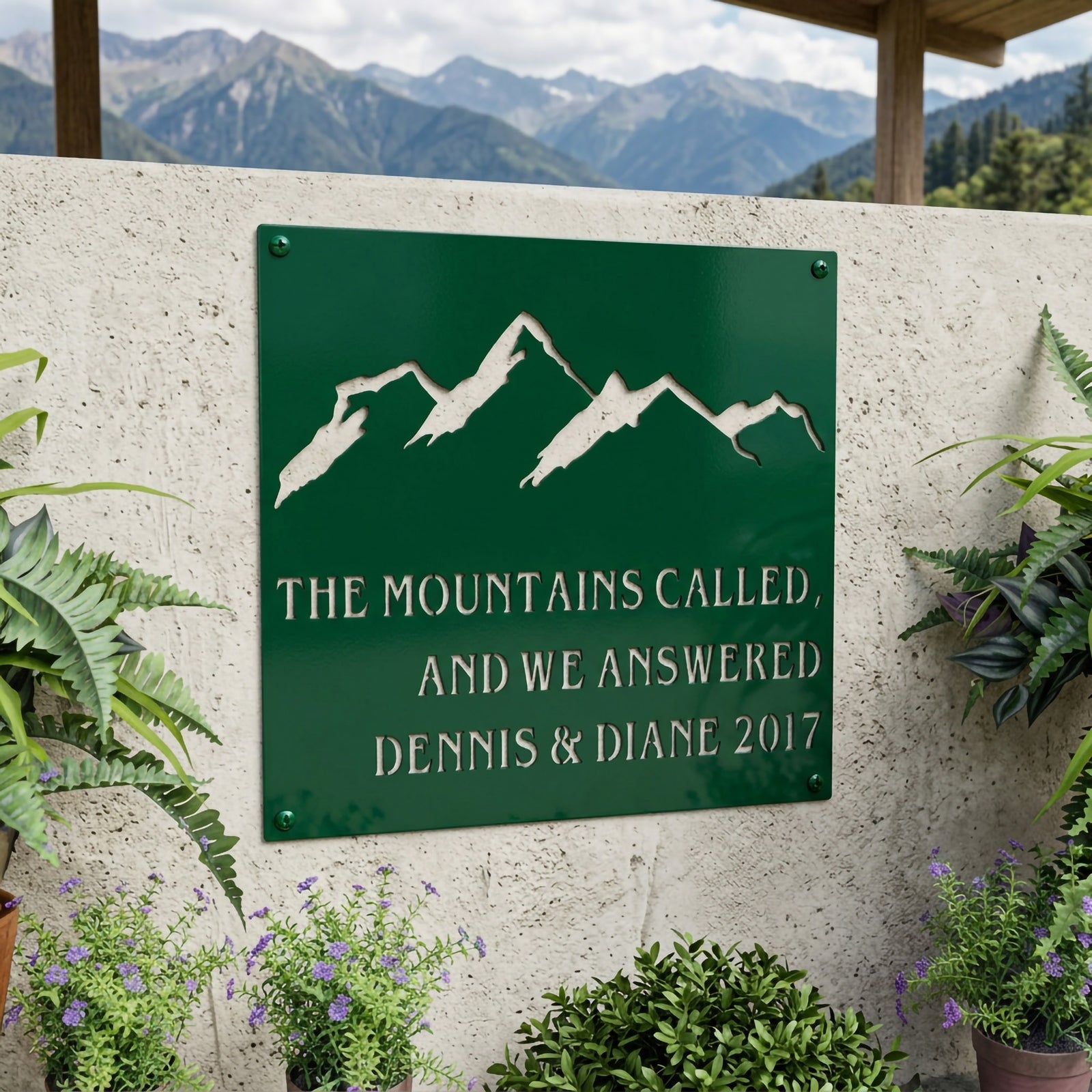 Emerald green mountain metal sign with personalized family names and established year displayed on an outdoor garden wall surrounded by plants and flowers.