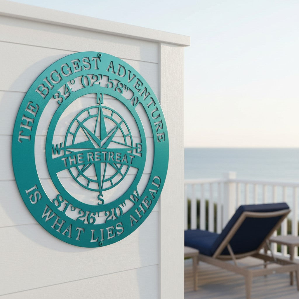 A circular metal compass sign finished in a bright teal color is mounted on a white wood-paneled wall. The outer ring features the phrase "THE BIGGEST ADVENTURE IS WHAT LIES AHEAD," while the inner circle displays custom coordinates and the text "THE RETREAT" across the compass rose. An outdoor deck with a blue lounge chair and the ocean are visible in the background.
