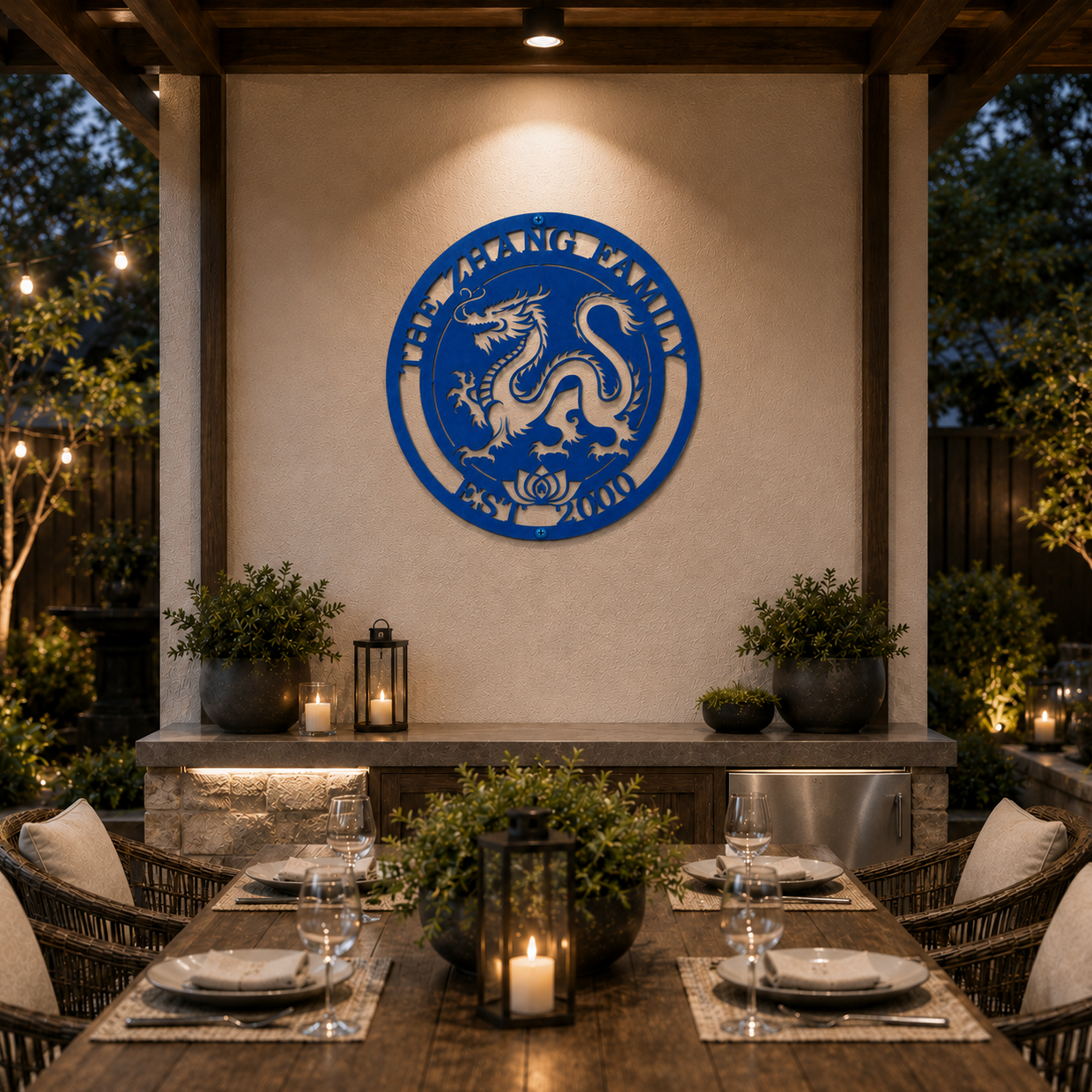 A vibrant cobalt blue circular metal sign featuring a laser-cut traditional Chinese dragon and a lotus flower at the base. The outer ring is customized with "THE ZHANG FAMILY EST. 2000." The sign is mounted on an outdoor stucco wall under a pergola, centered over an upscale patio dining table arranged with candles and wine glasses.