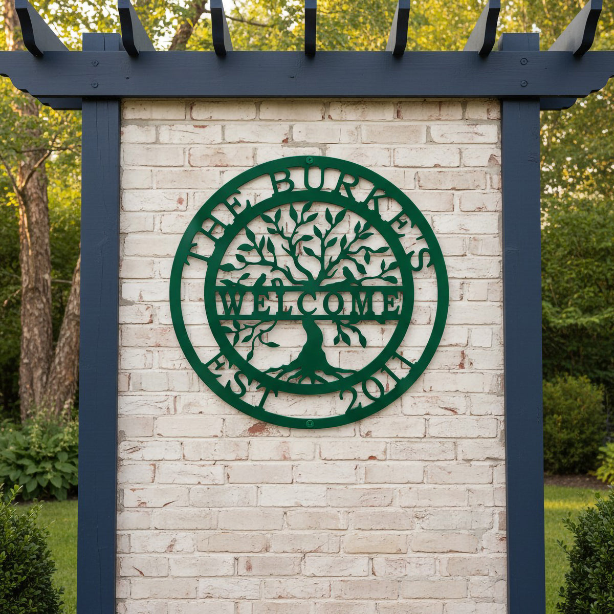 A dark green circular metal sign reading "The Burkes Welcome Est. 2011" with an intricate tree of life silhouette, mounted on a white brick wall under a dark blue pergola.