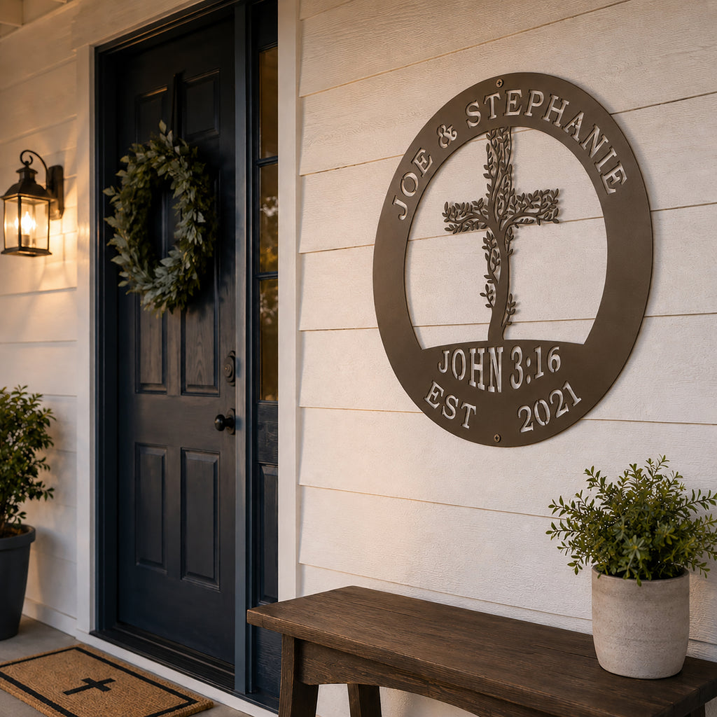 A circular metal family plaque with an oil rubbed bronze finish is mounted on a white clapboard exterior wall next to a dark blue front door. The sign displays the custom names "JOE & STEPHANIE" and "JOHN 3:16" around a central leafy cross. A wooden bench sits below the sign under a classic black entry lantern. 