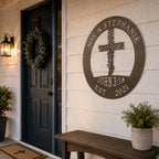 A circular metal family plaque with an oil rubbed bronze finish is mounted on a white clapboard exterior wall next to a dark blue front door. The sign displays the custom names "JOE & STEPHANIE" and "JOHN 3:16" around a central leafy cross. A wooden bench sits below the sign under a classic black entry lantern. 