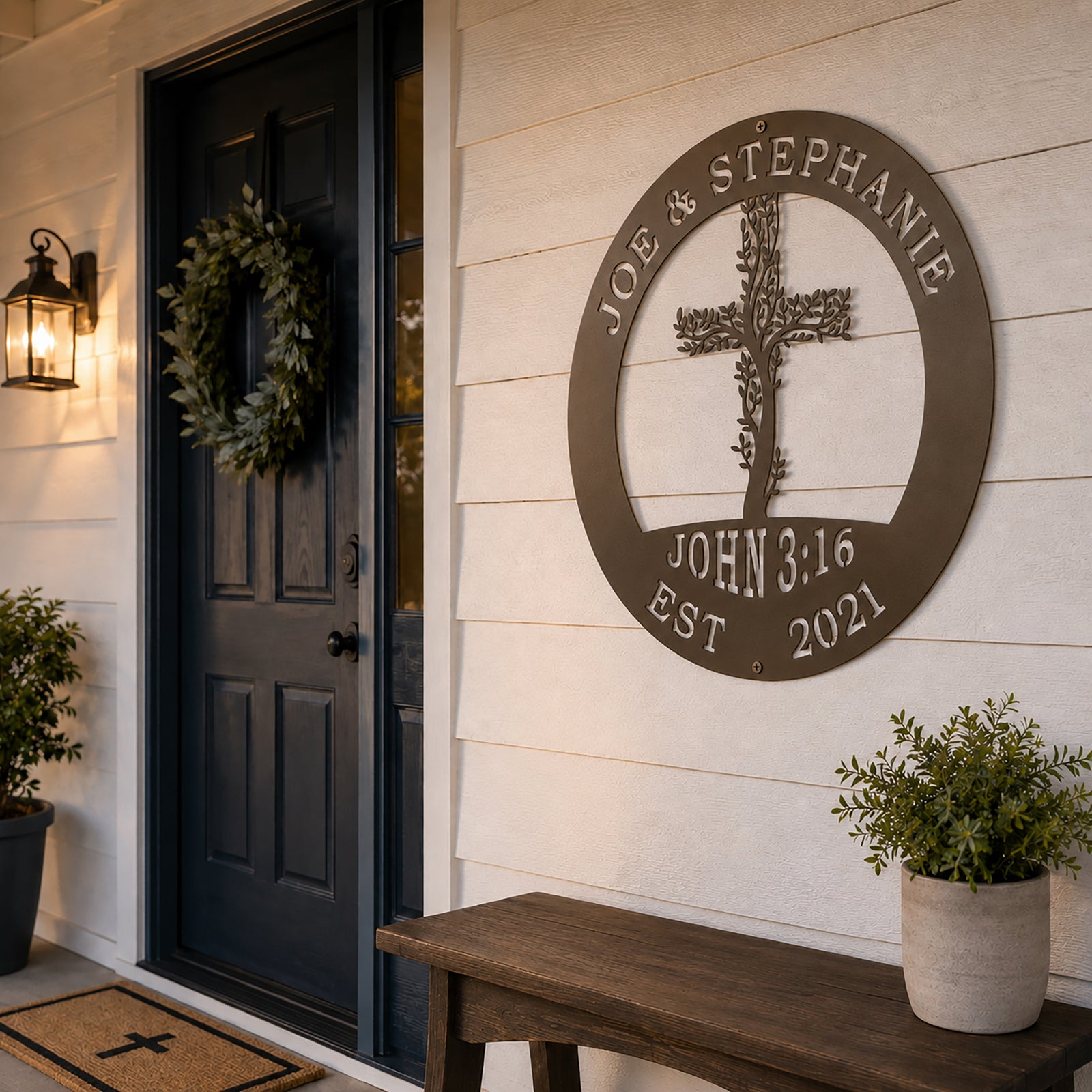 A circular metal family plaque with an oil rubbed bronze finish is mounted on a white clapboard exterior wall next to a dark blue front door. The sign displays the custom names "JOE & STEPHANIE" and "JOHN 3:16" around a central leafy cross. A wooden bench sits below the sign under a classic black entry lantern. 