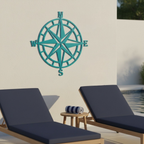  A teal-colored metal compass rose sign hanging on a white exterior wall above two wooden lounge chairs with dark blue cushions next to a swimming pool.