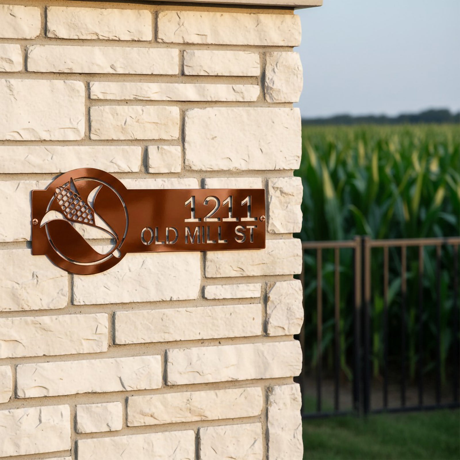 Tuscan copper corn address plaque with 1211 Old Mill St displayed on a stone entry pillar near a green cornfield