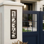 Dark bronze vertical metal house number sign 12345 mounted on a white front gate pillar beside a modern navy gate
