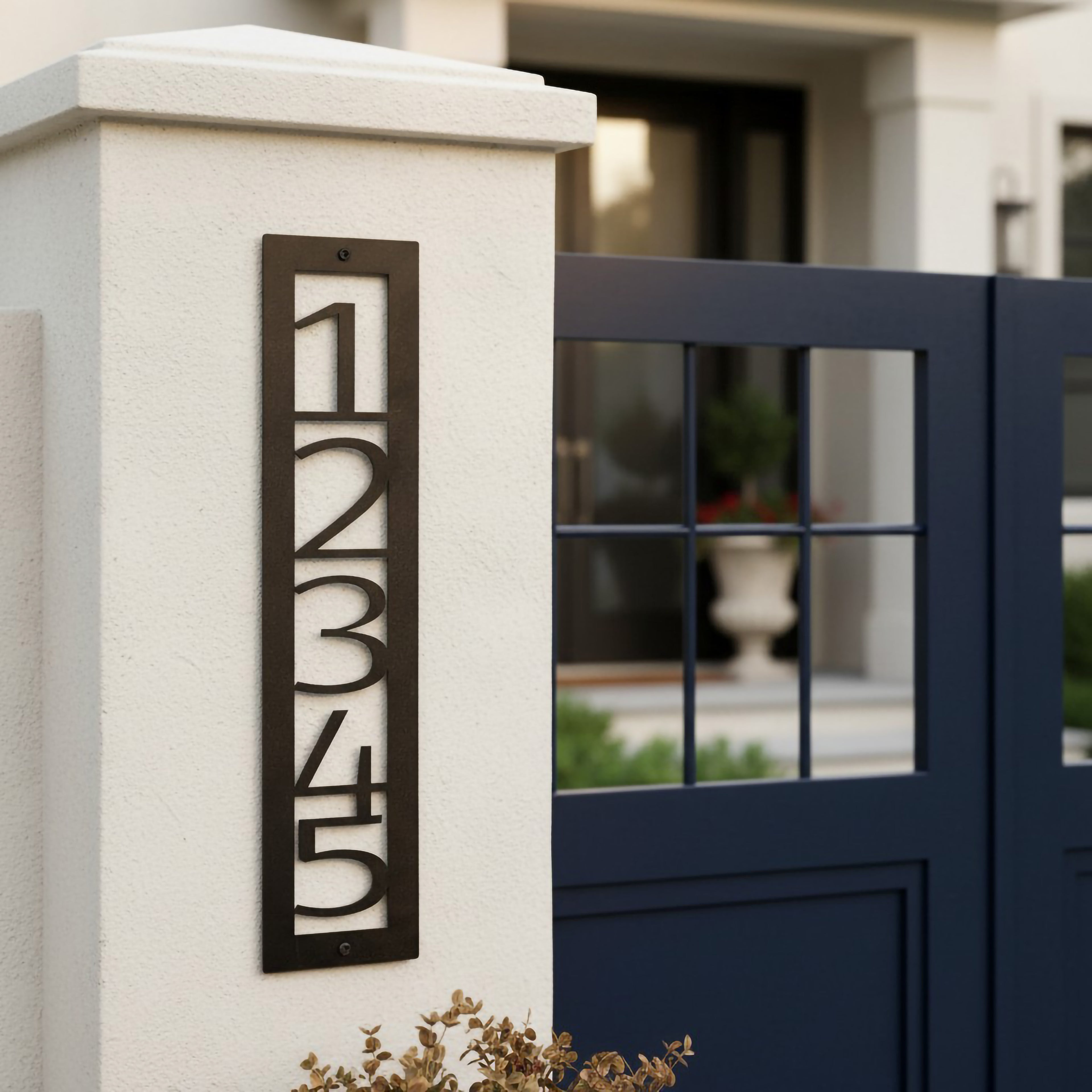 Dark bronze vertical metal house number sign 12345 mounted on a white front gate pillar beside a modern navy gate