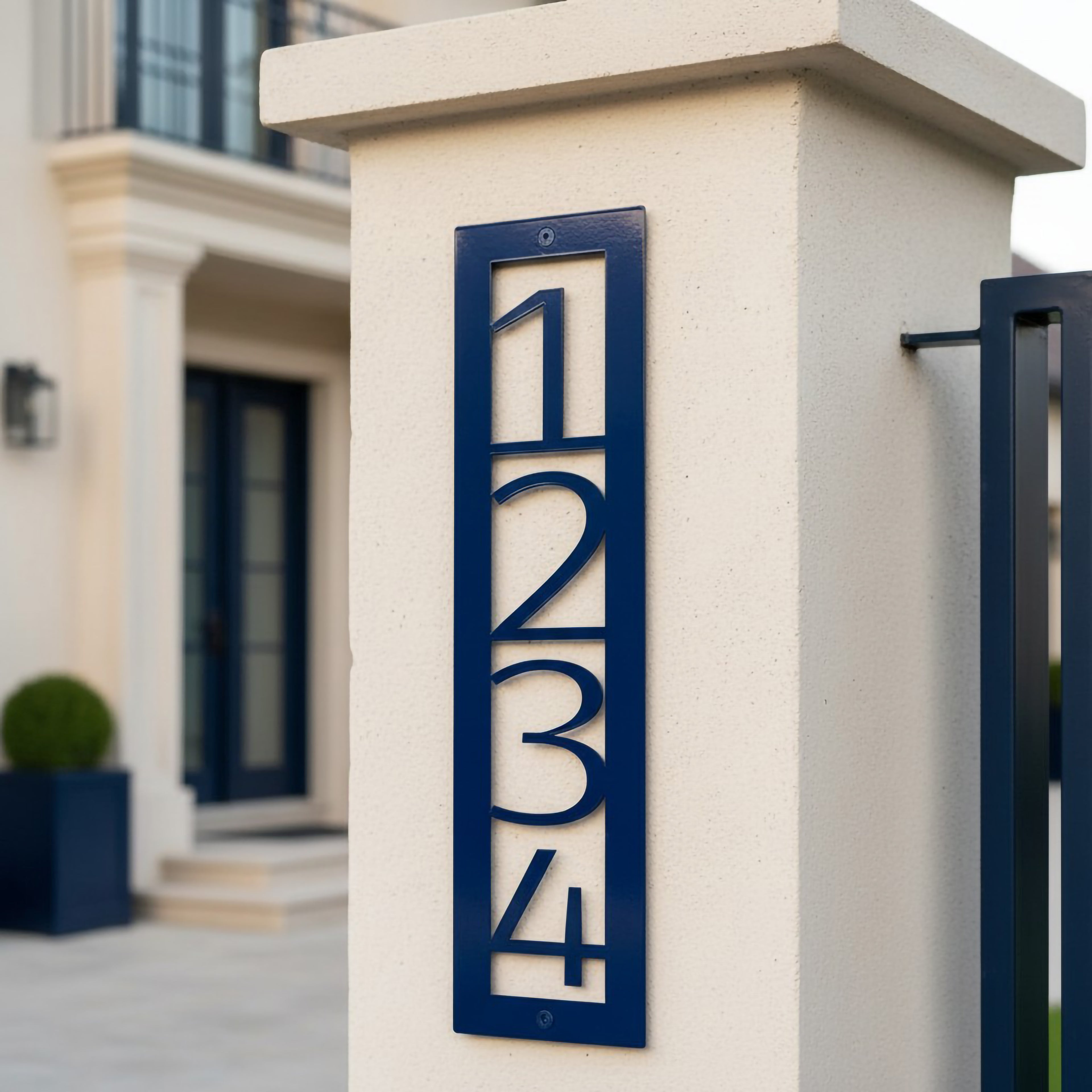 Navy blue vertical metal house number sign 1234 mounted on a white stucco pillar beside a modern entry gate