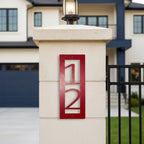 Red vertical metal house number sign 12 mounted on a white gate pillar in front of a modern home