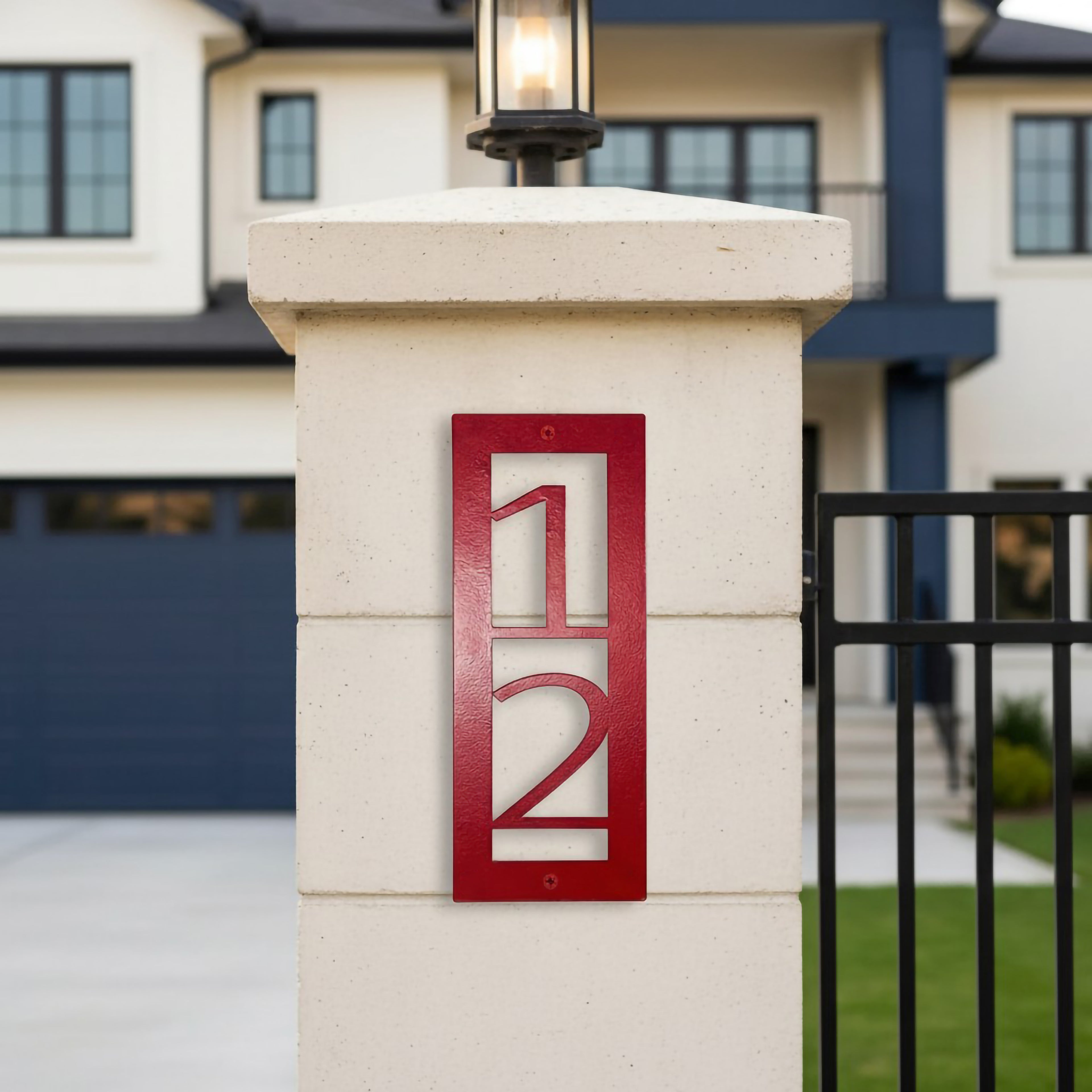 Red vertical metal house number sign 12 mounted on a white gate pillar in front of a modern home