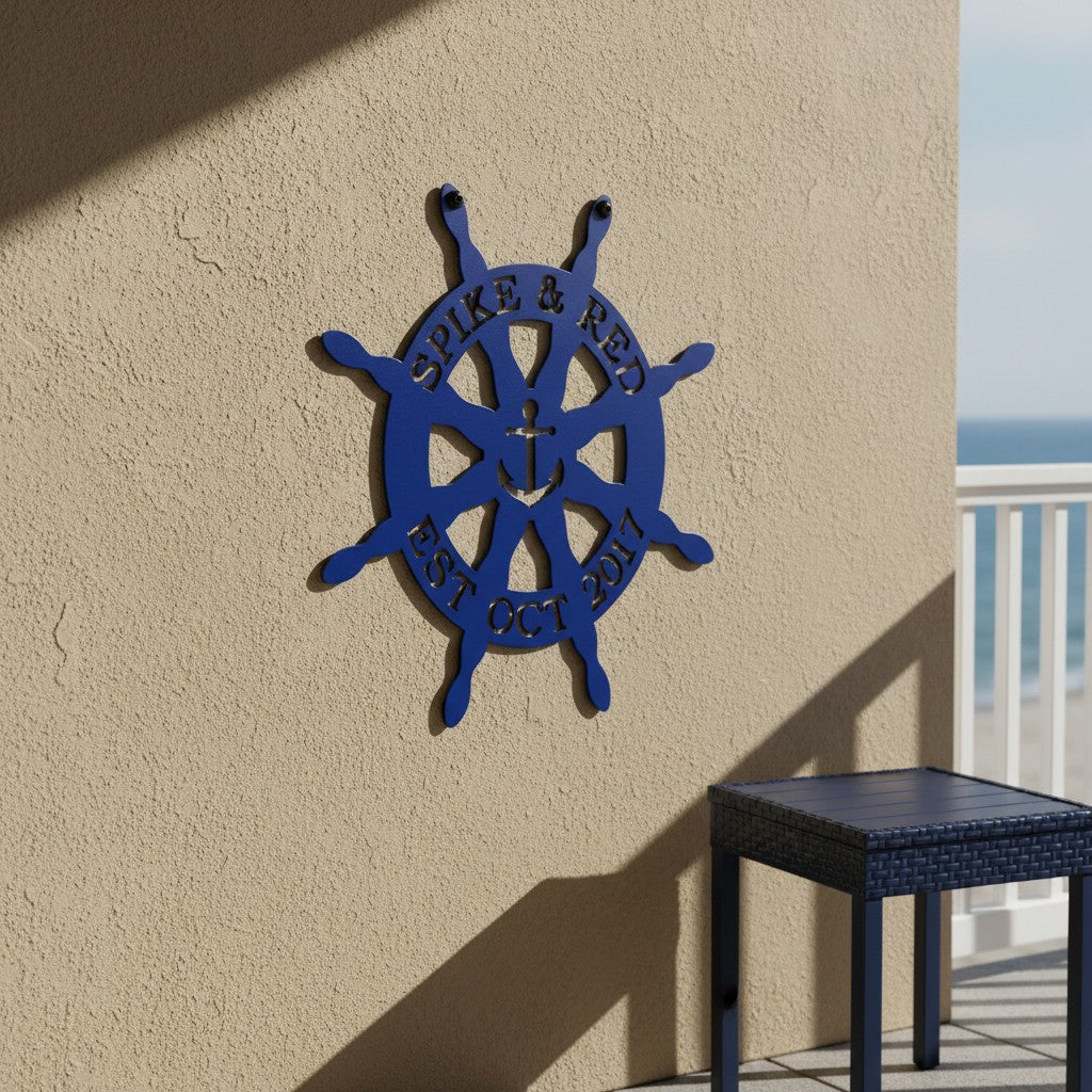 Royal blue custom metal ship wheel anchor sign displayed on a warm beige stucco exterior wall next to a dark blue wicker patio table.