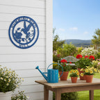 Textured navy blue custom metal round sign featuring a hummingbird silhouette layout, displayed centrally on an outdoor white shiplap wall next to a wooden table with potted red geraniums and a bright blue watering can.