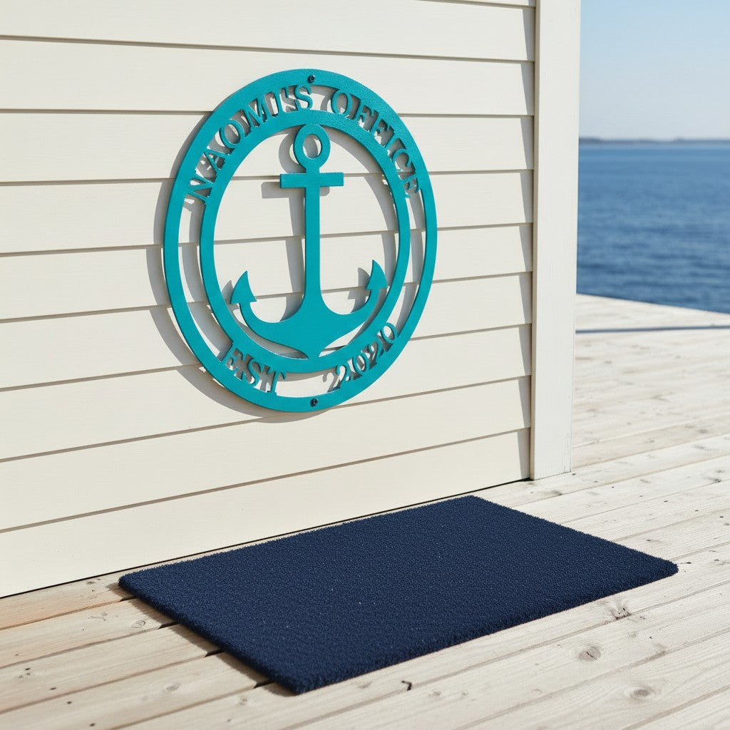 Bright teal custom metal anchor name sign displayed on a white horizontal siding wall directly above a navy blue doormat on a sunlit wooden deck.