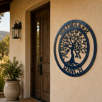 A circular metal sign finished in a deep navy blue color is mounted on a textured tan exterior wall near a dark wooden front door. The sign features an intricate Tree of Life cut-out design with the words "WAYMAKER" on the top rim and "RANCH" on the bottom rim. A rustic stone planter pot rests on the porch nearby.