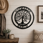 A textured satin black circular metal sign reading "WAYMAKER RANCH" around a central Tree of Life design is centered on a white shiplap entryway wall. A cowboy hat hangs from a coat rack on the left above a rustic wooden bench with a ceramic mug and potted greenery.