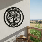 A matte black circular metal landscape sign featuring a laser-cut Tree of Life and "WAYMAKER RANCH" lettering is fixed to an outdoor white wall. Below the sign sits a small wooden side table holding a straw cowboy hat and work gloves, set next to a wooden balcony railing overlooking fields.