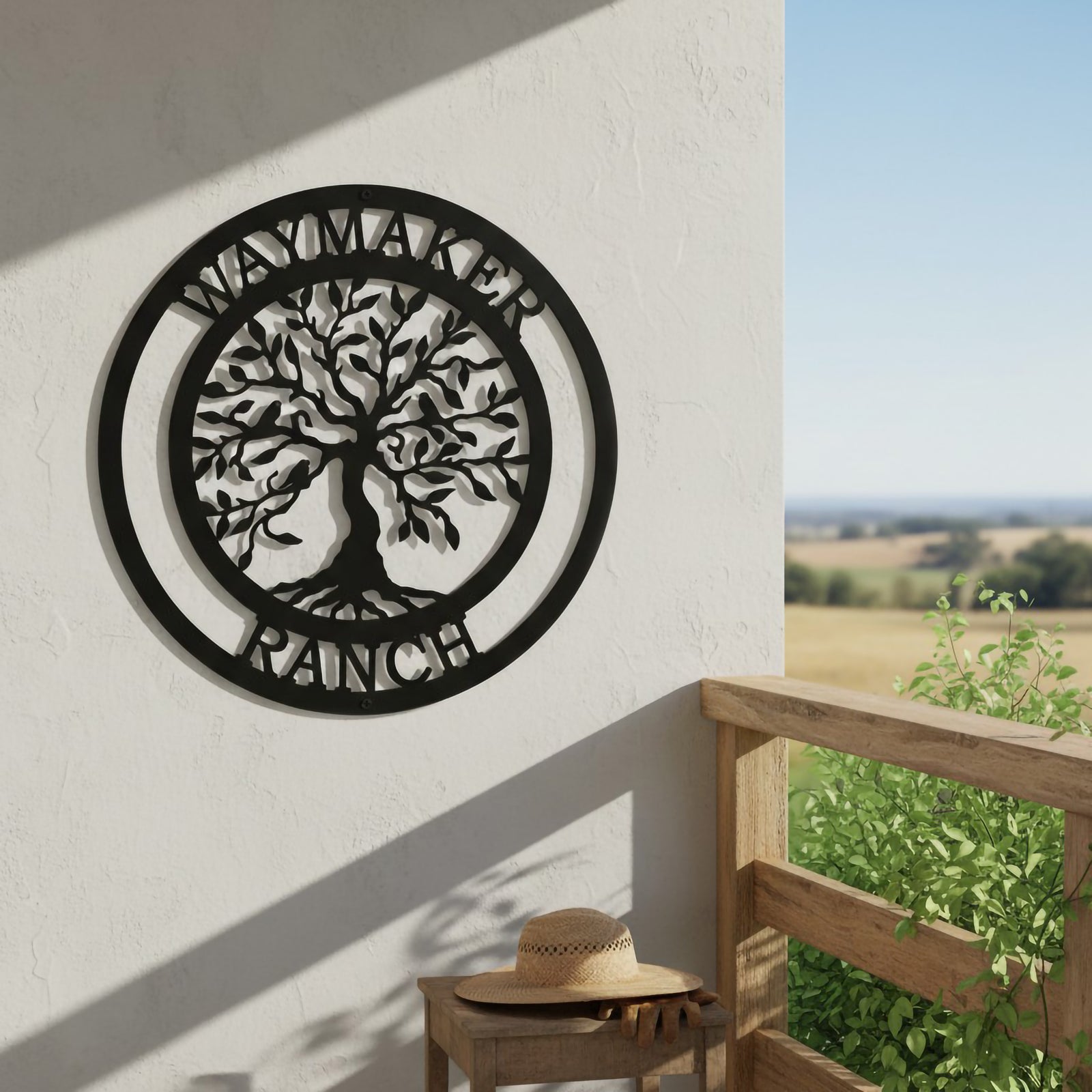 A matte black circular metal landscape sign featuring a laser-cut Tree of Life and "WAYMAKER RANCH" lettering is fixed to an outdoor white wall. Below the sign sits a small wooden side table holding a straw cowboy hat and work gloves, set next to a wooden balcony railing overlooking fields.
