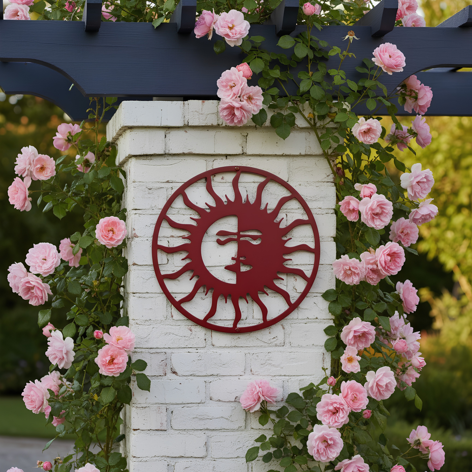 A dark red circular metal sun face sign attached to a white brick pillar, beautifully framed by climbing pink roses growing along a dark blue wooden arbor.