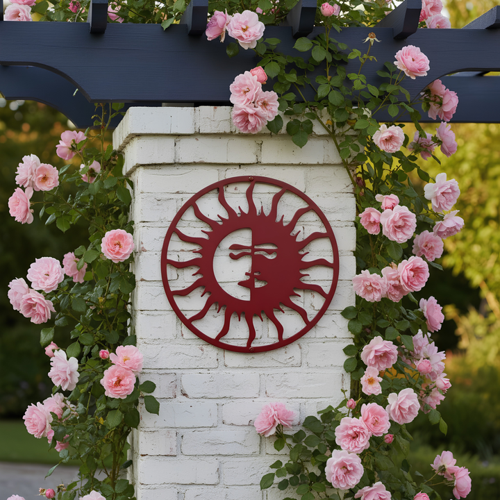 A dark red circular metal sun face sign attached to a white brick pillar, beautifully framed by climbing pink roses growing along a dark blue wooden arbor.