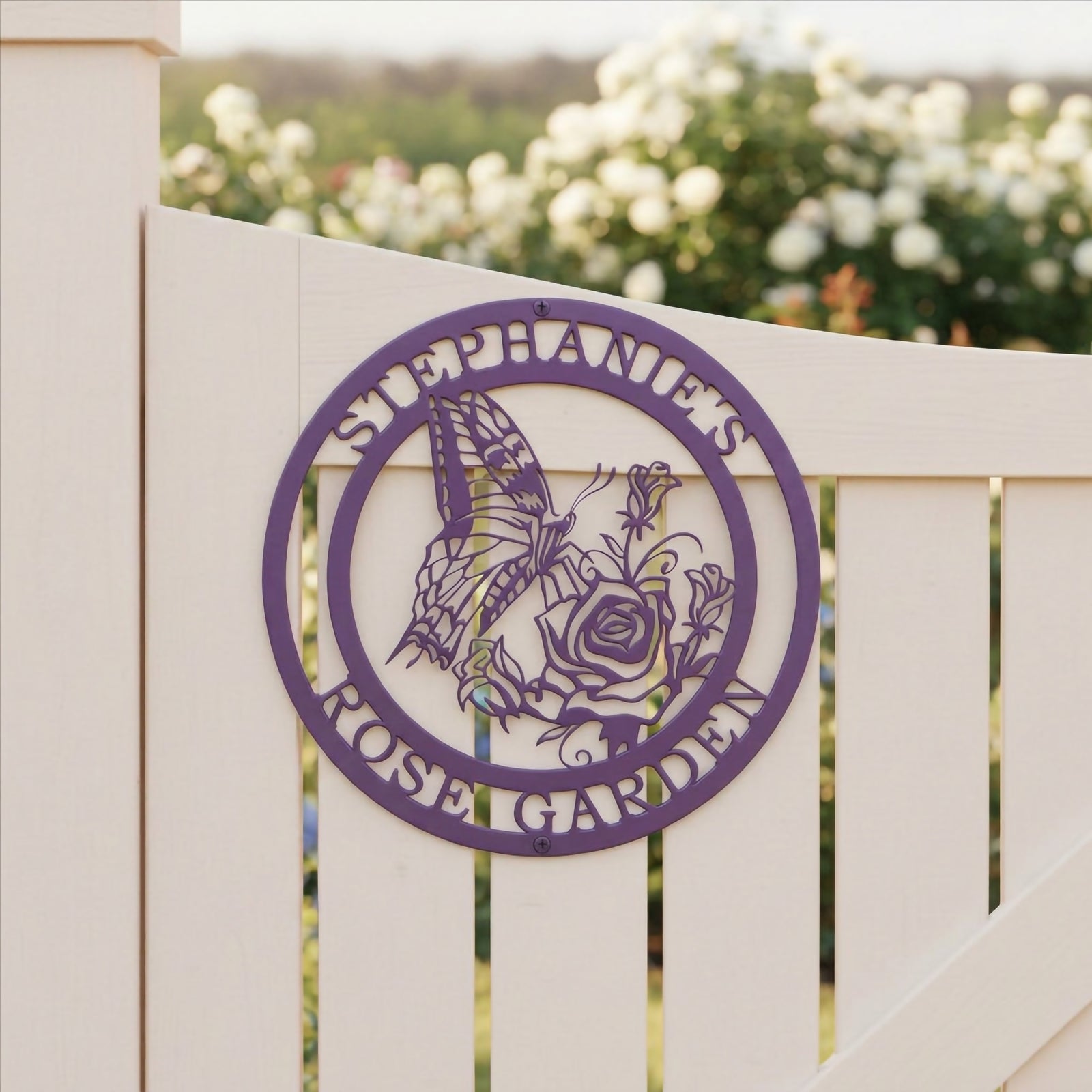A purple circular metal sign featuring a butterfly on a rose cutout, reading "Stephanie's Rose Garden" and attached to a white slatted garden gate.