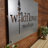 RM Custom Size Test metal business logo sign displayed on a wooden wall, accompanied by a potted houseplant, representing professional branding for office or company settings.