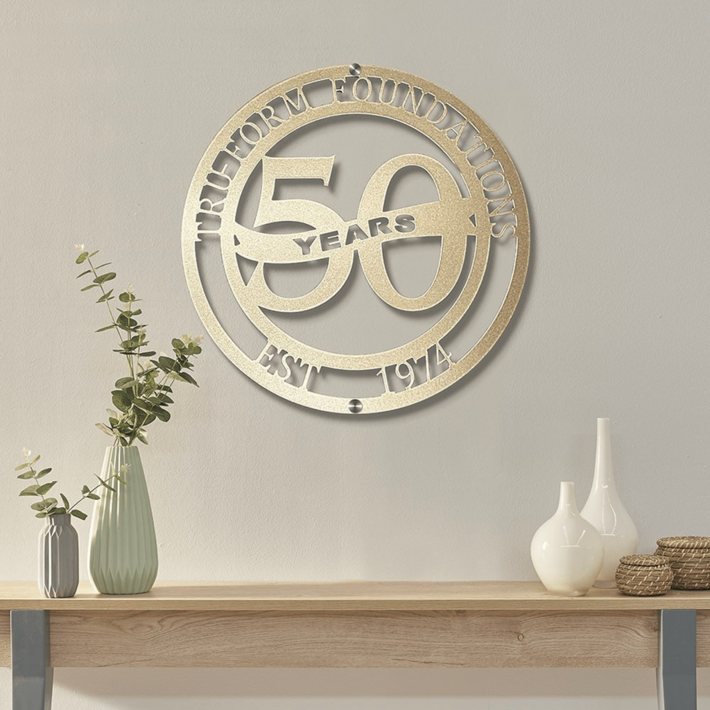 a decorative metal sign hanging on a wall, celebrating the 50th anniversary of a foundation. The sign is surrounded by a circular arrangement of decorative elements, including a vase with greenery and two white vases.