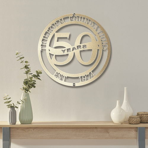 a decorative metal sign hanging on a wall, celebrating the 50th anniversary of a foundation. The sign is surrounded by a circular arrangement of decorative elements, including a vase with greenery and two white vases.