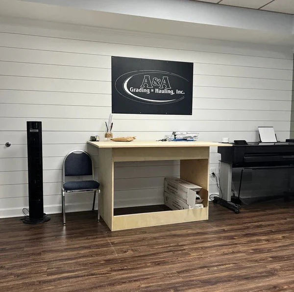 Wall-mounted black sign says “A&A Grading & Hauling, Inc.” in bold letters above a wooden desk against horizontal paneled walls.