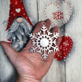 Hand holding a white snowflake ornament in front of a festive holiday background featuring a decorative gnome, red heart accents, and green seasonal greenery, creating a cozy and whimsical Christmas scene.
