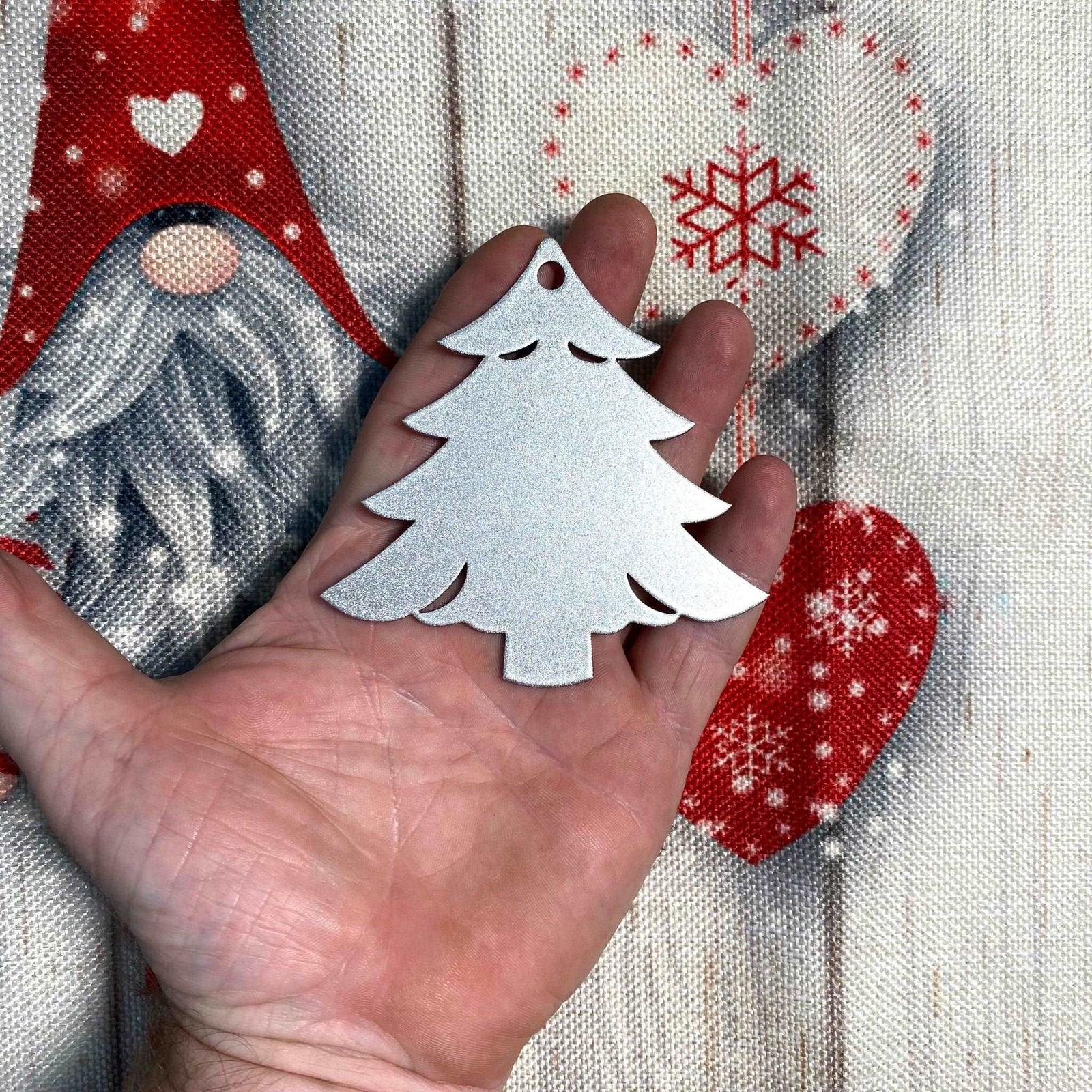 Hand holding a silver Christmas tree ornament against a festive holiday background with green branches, twinkling lights, and seasonal decorations, creating a warm and cheerful Christmas atmosphere.
