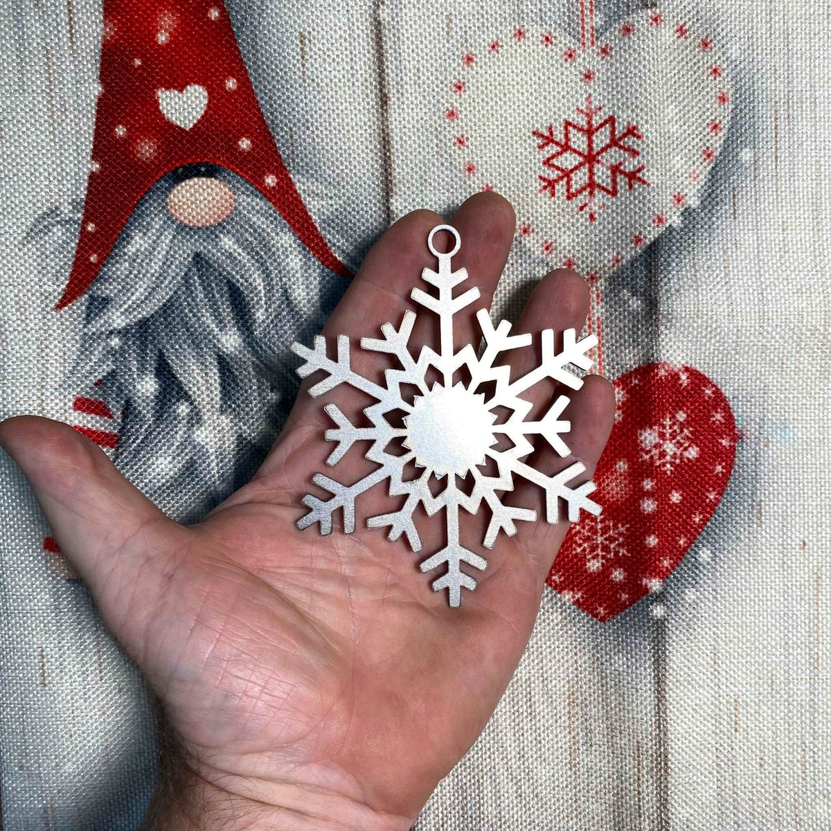 Hand holding a white snowflake Christmas ornament with festive background featuring a holiday gnome and red hearts, seasonal decoration for Christmas and winter décor