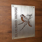 Metal sign featuring the text “Bluebird House” alongside a bluebird illustration, mounted on a wooden background, highlighting rustic charm and vibrant decorative design.