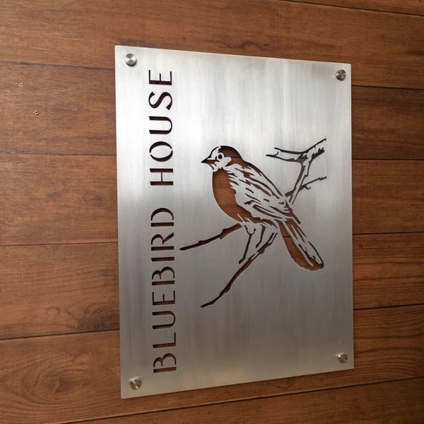 Metal sign featuring the text “Bluebird House” alongside a bluebird illustration, mounted on a wooden background, highlighting rustic charm and vibrant decorative design.