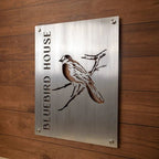 Metal sign featuring the text “Bluebird House” alongside a bluebird illustration, displayed on a wooden background, highlighting rustic charm and vibrant, decorative design.