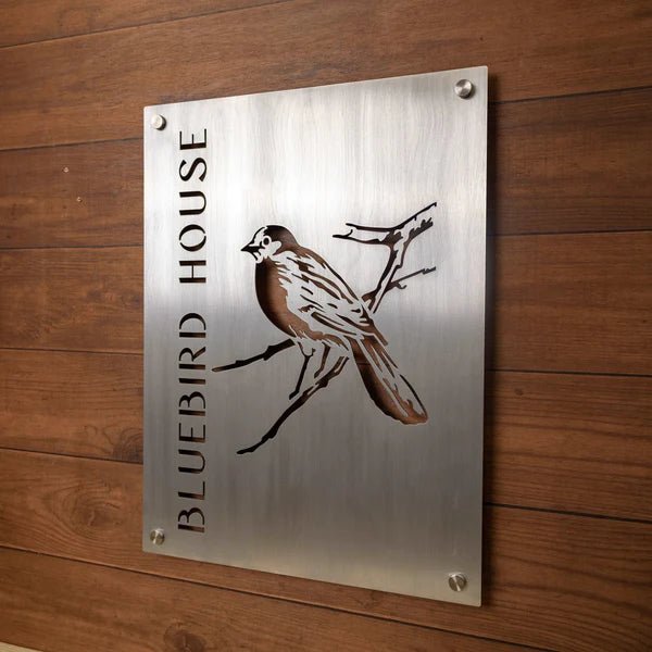 Metal sign featuring the text “Bluebird House” alongside a bluebird illustration, displayed on a wooden background, highlighting rustic charm and vibrant, decorative design.