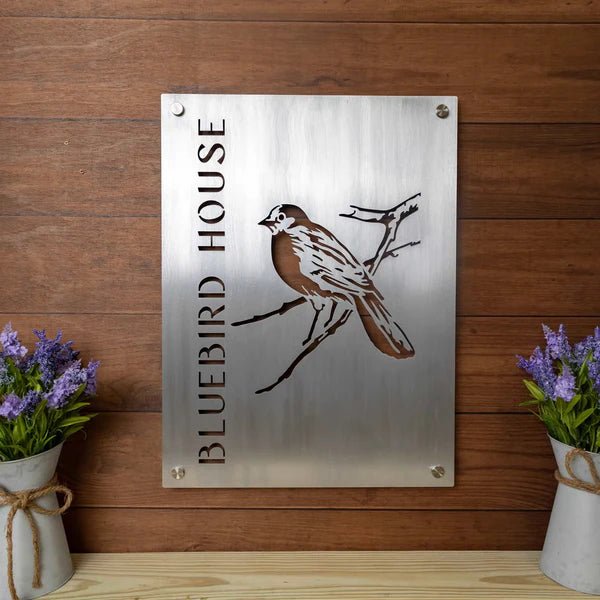 Metal sign displaying “Bluebird House” with a bluebird illustration on a wooden background, showcasing rustic style, vibrant colors, and durable craftsmanship.