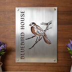 Metal sign featuring a bluebird illustration and the text “Bluebird House,” displayed on a wooden background, showcasing vibrant colors and rustic decorative design.