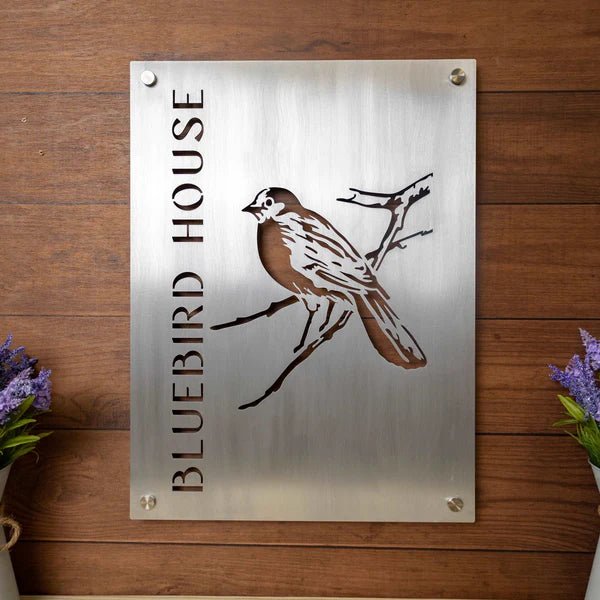 Metal sign featuring a bluebird illustration and the text “Bluebird House,” displayed on a wooden background, showcasing vibrant colors and rustic decorative design.